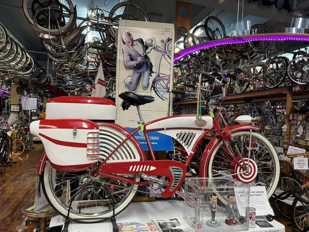 A red-and-white Pee-wee Herman movie-prop cruiser displayed beneath a Pee-wee poster, surrounded by walls of vintage bicycles
